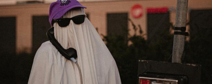 A person in a white sheet, representing a ghost, wearing a purple hat that says spooky. On a payphone outside in the street