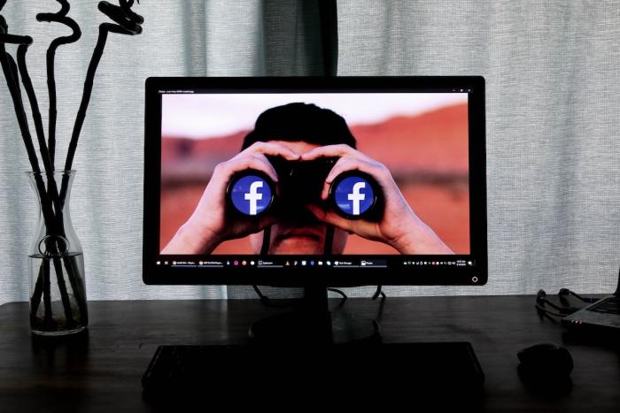 screen with individual holding binoculars with facebook sign on end