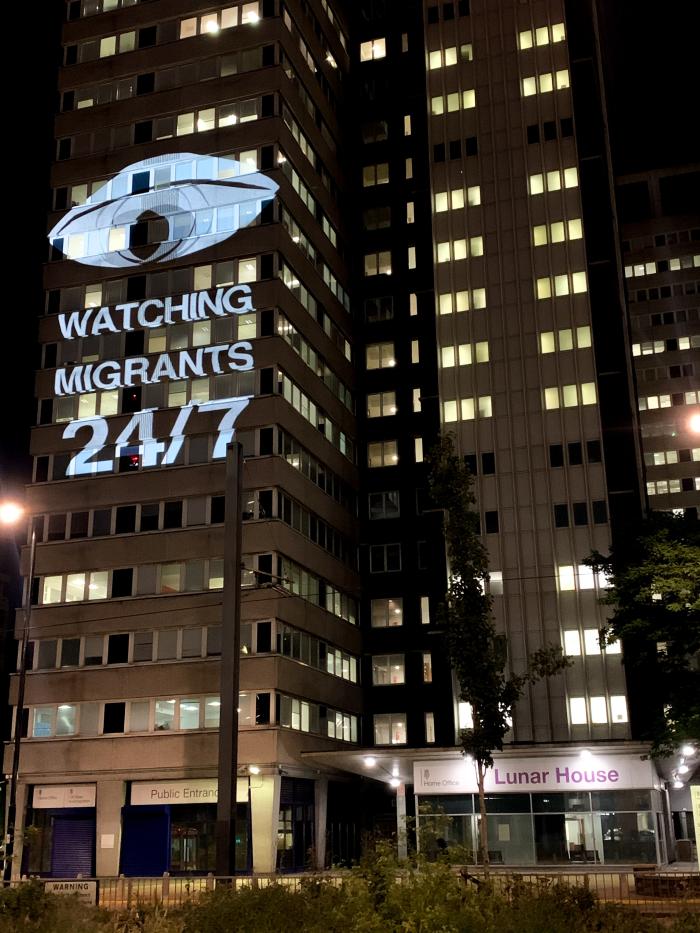 Light projection on UK Home Office building reading "WATCHING MIGRANTS 24/7"