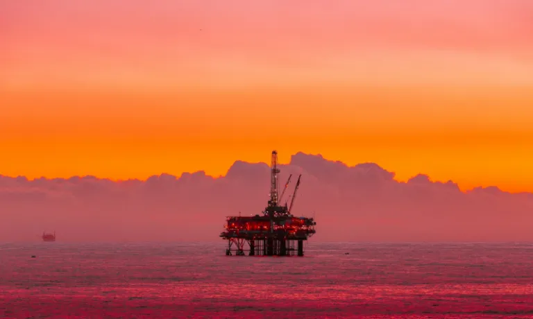 An offshore oil platform stands in the sea at sunset, framed by orange and pink skies, evoking the role of fossil fuel industries in debates about public subsidy, risk, and long-term harm.