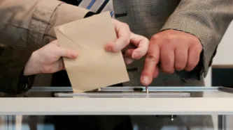Image of three hands voting, casting a ballot paper into a box together.